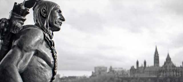 bronze statue of traditionally dressed Aboriginal man with braids with Canada's Parliament Buildings in background